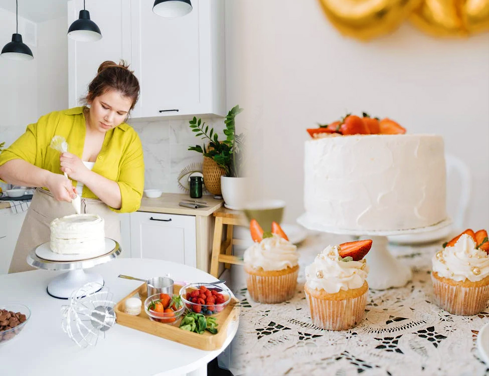 A woman is frosting a cake in a kitchen, and some frosted cakes are displayed on a table.