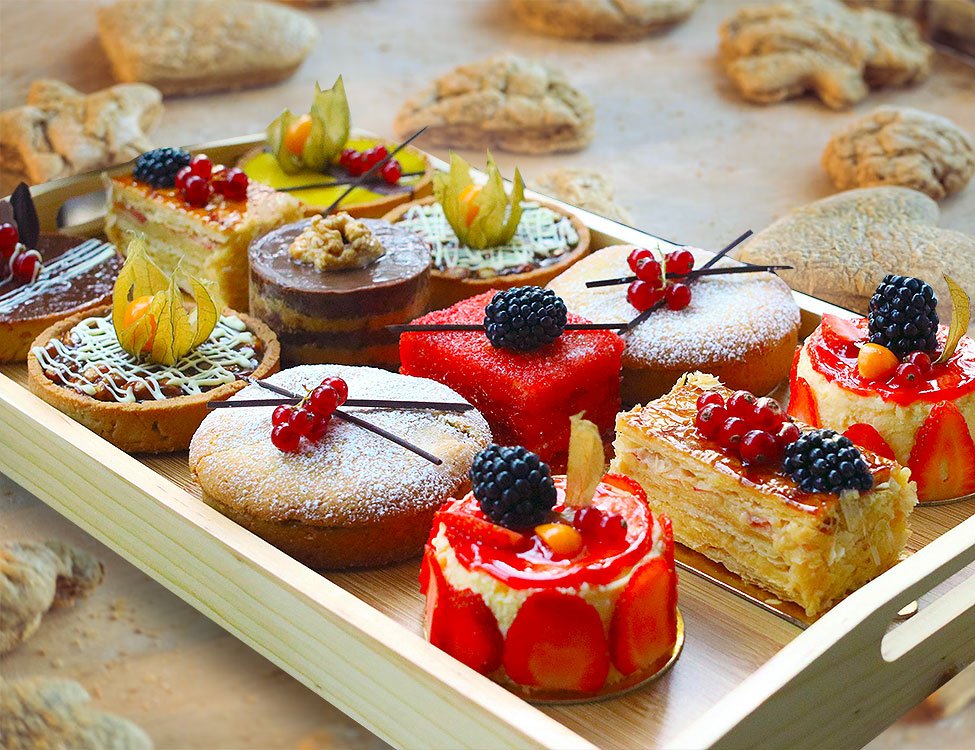 A wooden tray of a variety of single-serving cakes decorated with berries, superimposed on a background of cookies on a tray